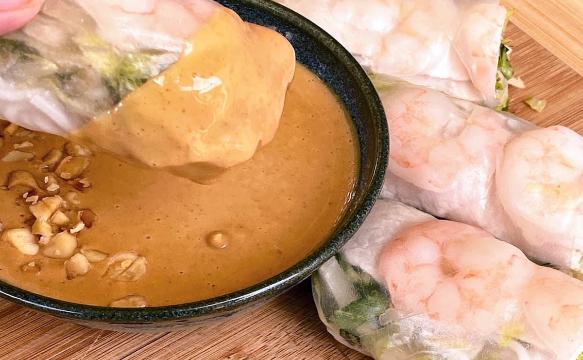 Fresh roll dipping into small bowl of peanut sauce. More fresh rolls at the foot of the bowl.
