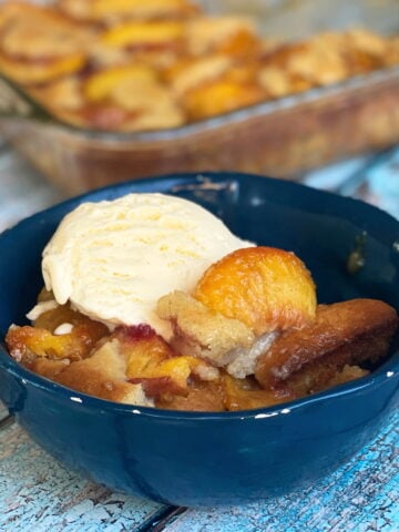 Peach cobbler and vanilla ice cream in a dessert bowl, with a pan of cobbler in the background.