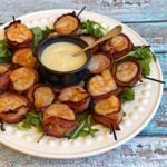 Plate of broiled, cooked bacon wrapped shrimps on a bed of arugula, with a small cup of pineapple-horseradish sauce in the center.
