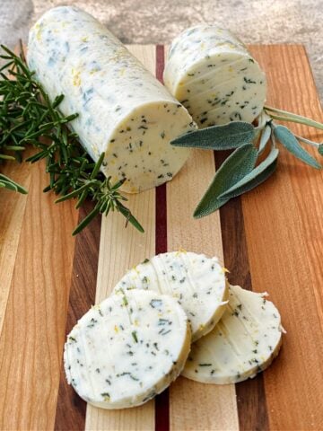 Compound butter roll and coins resting on small cutting board, garnished with fresh sage and rosemary.