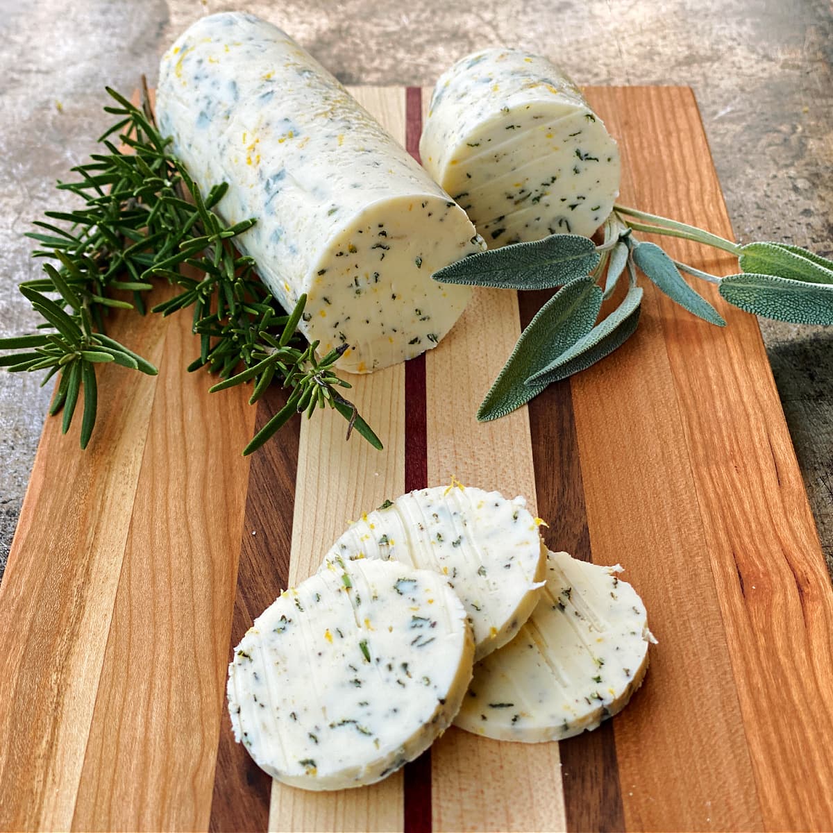 Compound butter roll and coins resting on small cutting board, garnished with fresh sage and rosemary. 