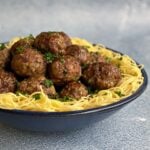 Bowl of cooked spaghetti noodles with a pile of baked meatballs in the center, garnished with chopped parsley.