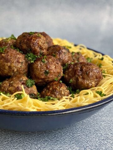 Bowl of cooked spaghetti noodles with a pile of baked meatballs in the center, garnished with chopped parsley.
