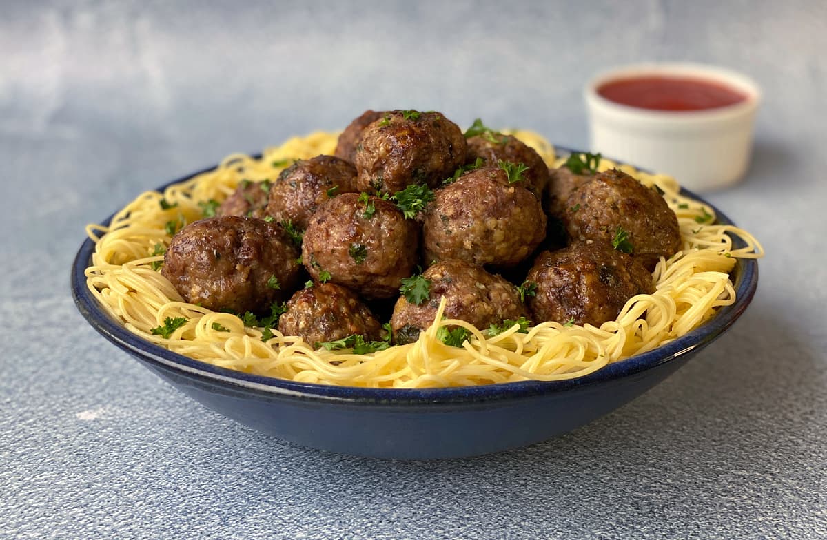 Bowl of cooked spaghetti noodles with a pile of baked meatballs in the center, garnished with chopped parsley. Spaghetti sauce in a ramekin in the background.