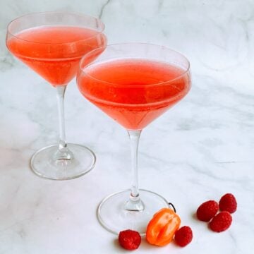 Two pink cocktails in martini glasses, with five raspberries and a habanero pepper at the base of one glass.