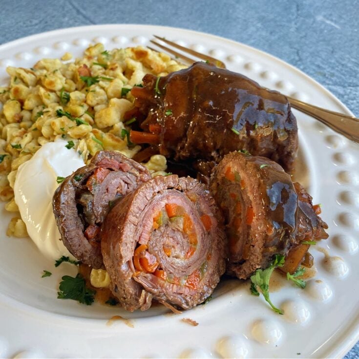 Two German Beef Rouladen rolls sliced on plate to show layered pinwheel, with sour cream and spätzle on the side.