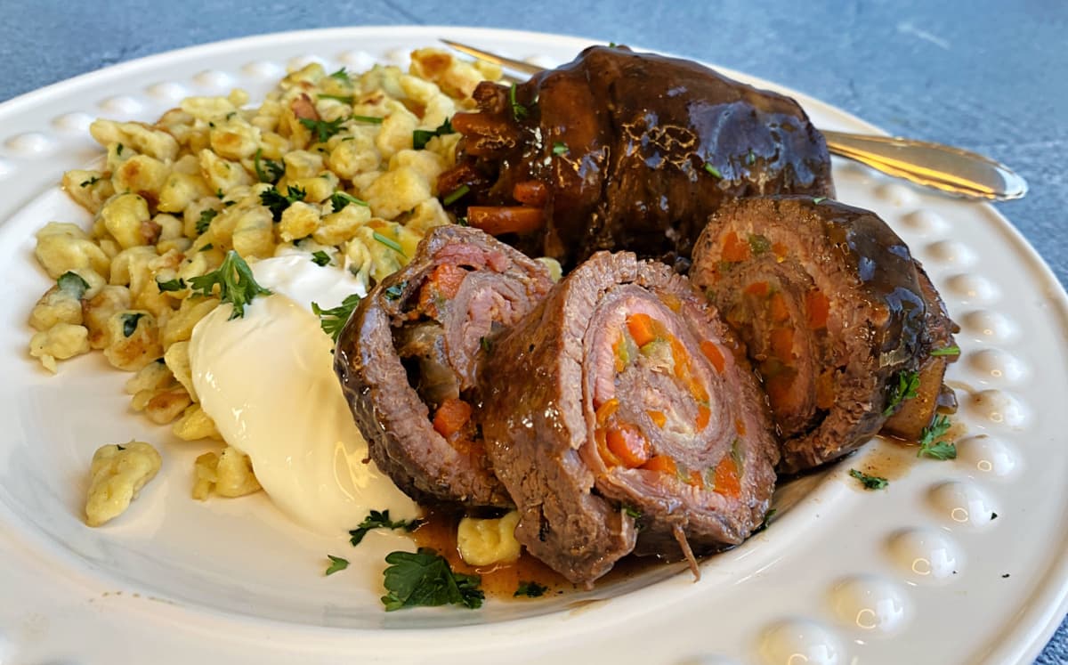 Two German Beef Rouladen rolls sliced on plate to show layered pinwheel, with sour cream and spätzle on the side.