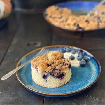 A round-cut piece of blueberry buckle on a plate, with a dollap of yogurt and a sprinkle of fresh blueberries on the side. The remaining unserved buckle is in the background.