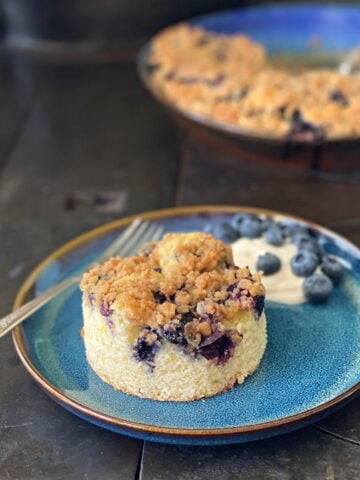 A round-cut piece of blueberry buckle on a plate, with a dollap of yogurt and a sprinkle of fresh blueberries on the side. The remaining unserved buckle is in the background.