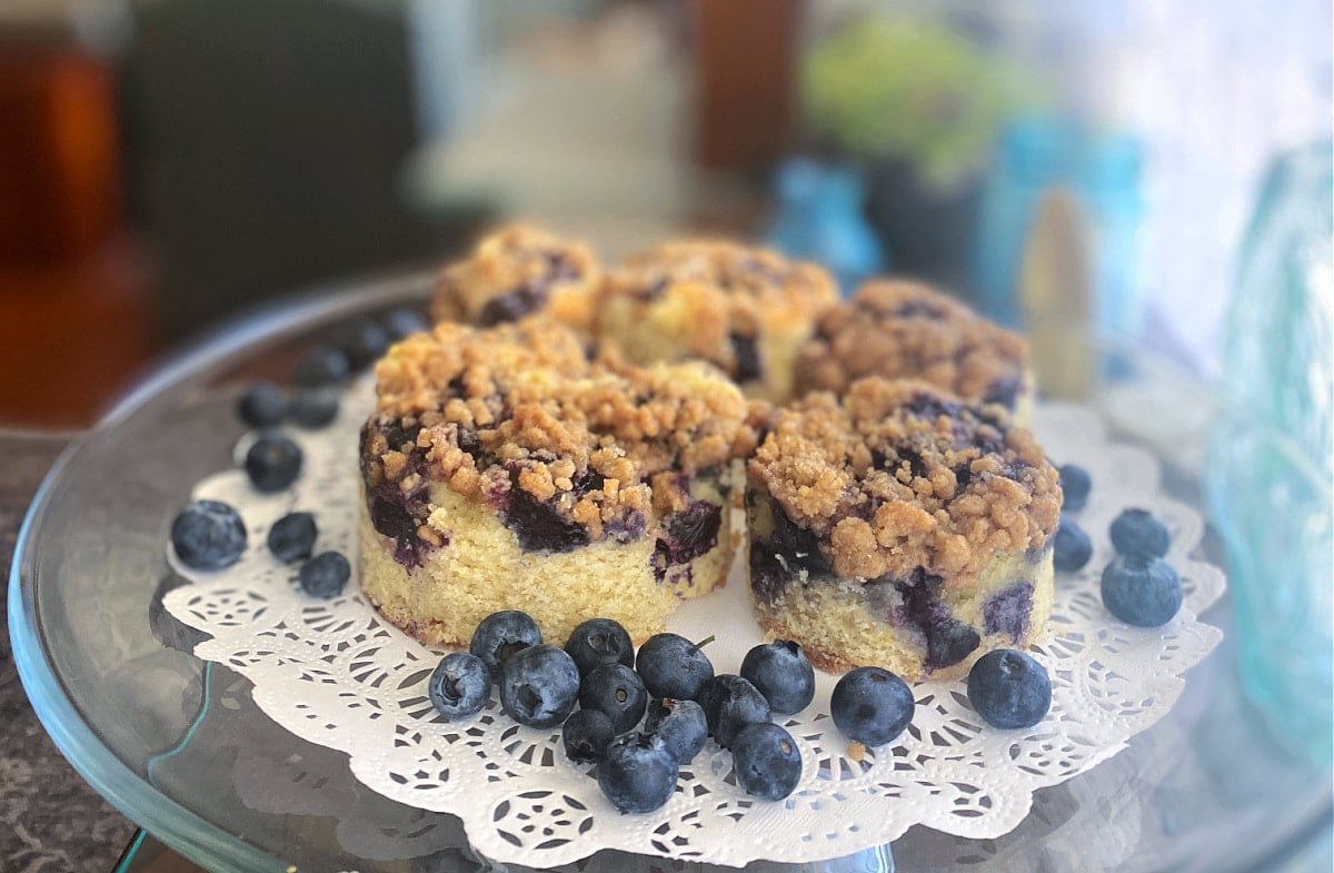Cake plate with a doily covered with five round blueberry buckle servings, cut using biscuit cutters of various sizes. 