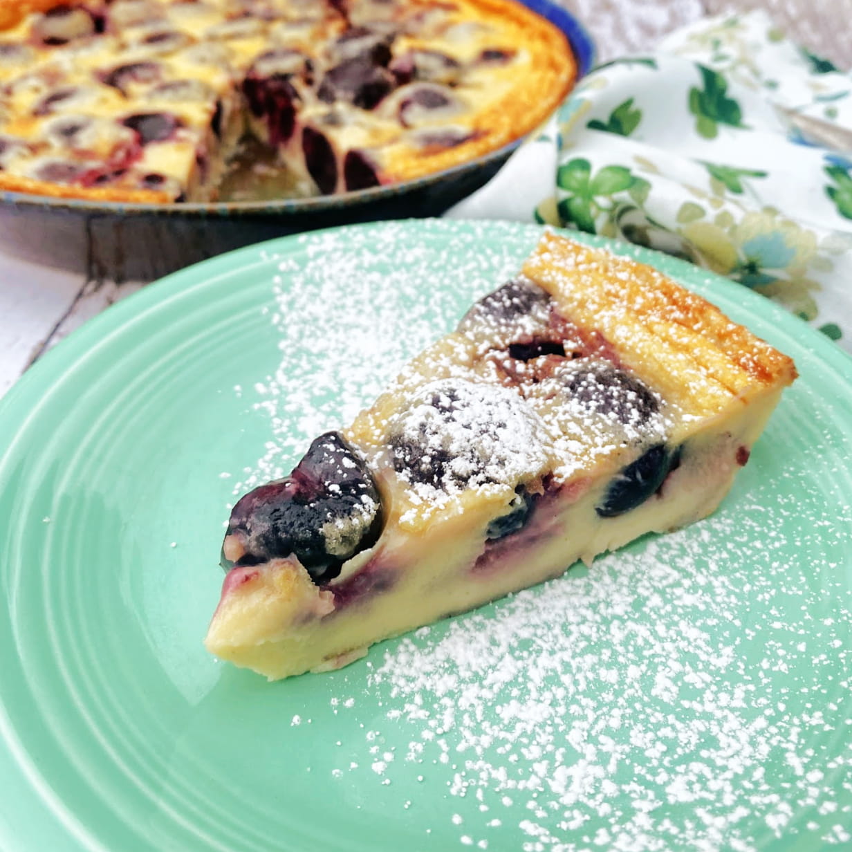 Slice of clafoutis on dessert plate. Pie plate with remaining clafoutis in background.