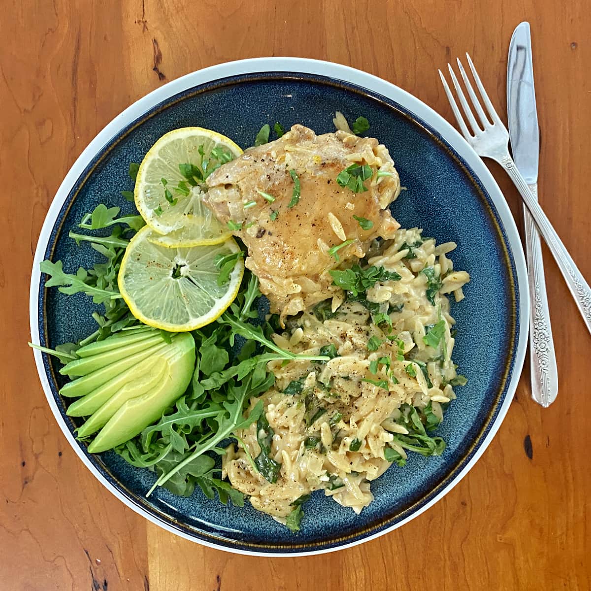 Overhead shot of plated lemon chicken thighs with creamy orzo, garnished with baby arugula, lemon slices, and avocado fan.