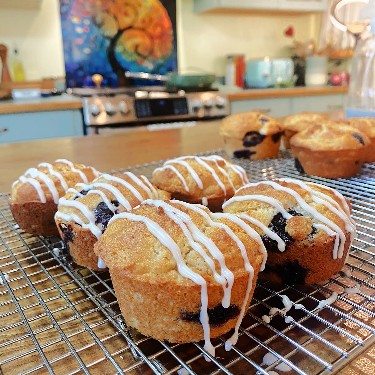 Baked lemon blueberry muffins on a cooling rack, lightly drizzled with icing. Vintage kitchen in the background. 