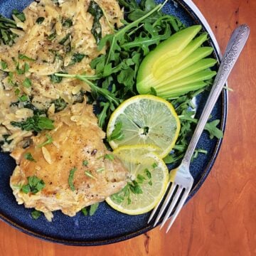 Overhead shot of plated lemon chicken thighs with creamy orzo, garnished with baby arugula, lemon slices, and avocado fan.