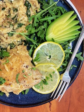 Overhead shot of plated lemon chicken thighs with creamy orzo, garnished with baby arugula, lemon slices, and avocado fan.