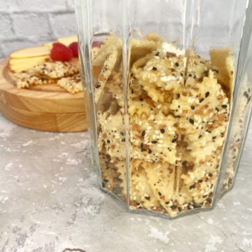 Close-up of rustic sourdough crackers stacked in an antique jar, with a cheese board of cheddar and raspberries in the background.