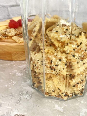 Close-up of rustic sourdough crackers stacked in an antique jar, with a cheese board of cheddar and raspberries in the background.