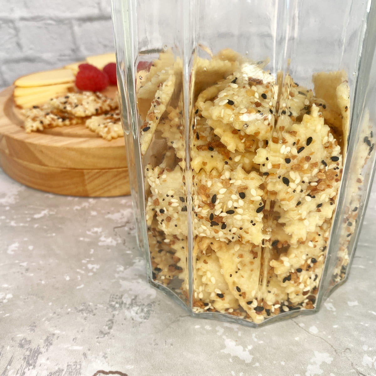 Close-up of rustic sourdough crackers stacked in an antique jar, with a cheese board of cheddar and raspberries in the background.