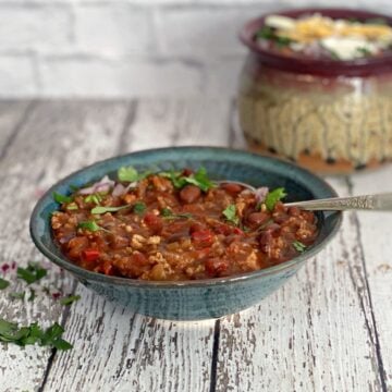 A hearty bowl of turkey chili topped with sour cream, cilantro, and red onion, with a mug of chili in the background.