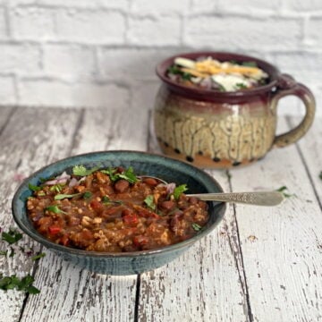 A hearty bowl of turkey chili topped with sour cream, cilantro, and red onion, with a mug of chili in the background.