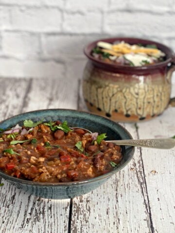A hearty bowl of turkey chili topped with sour cream, cilantro, and red onion, with a mug of chili in the background.