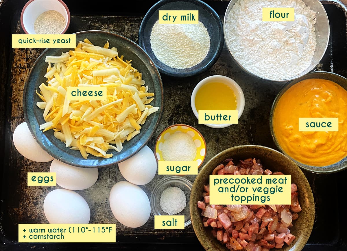 Ingredients for cast iron breakfast pizza including flour, yeast, eggs, cheese, ham, bacon, and vegetables, arranged on an old sheet pan.