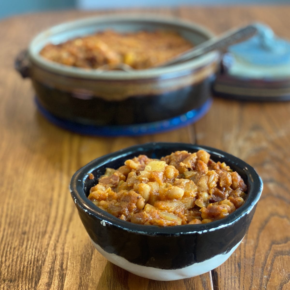 Homemade pineapple baked beans with bacon in a small bowl, with a pot of baked beans in the background.
