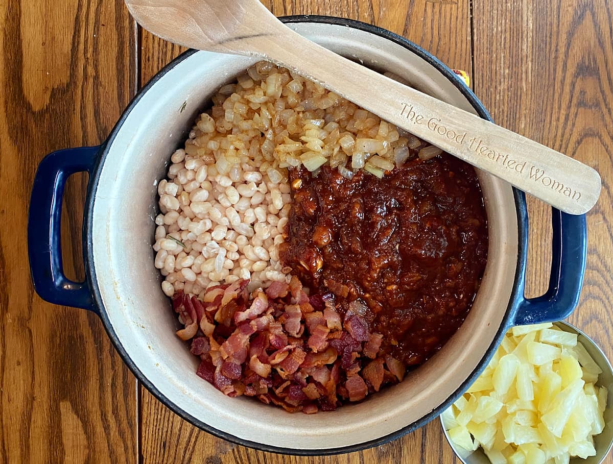 Beans, sauce, sauteed onions, and bacon in a Dutch oven, with pineapple tidbits in a small bowl ot the side.